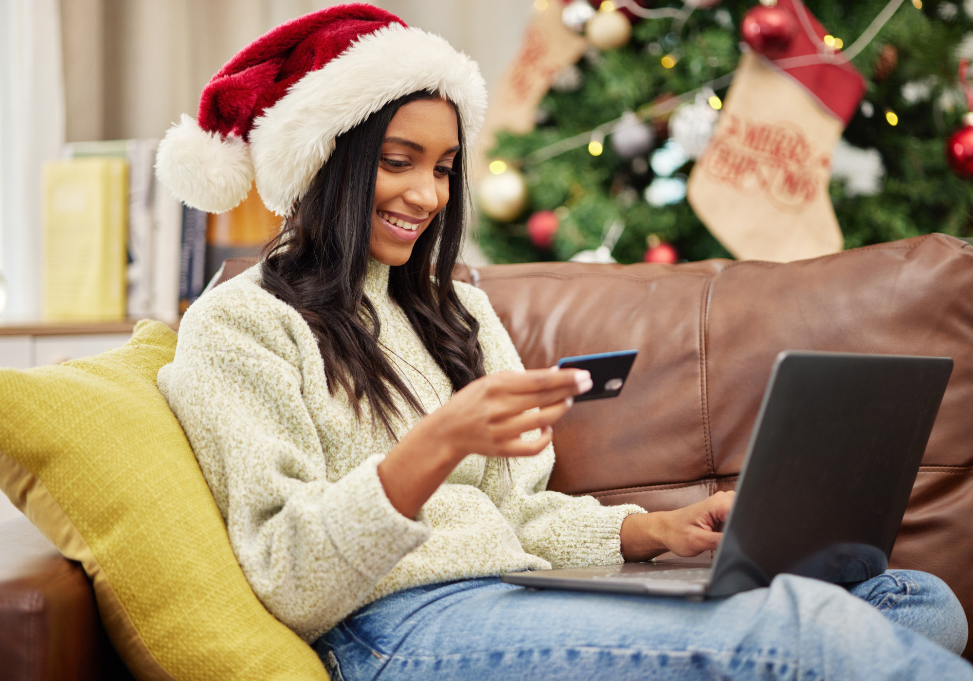 Woman with Santa hat sitting on sofa holding The Police Credit Union credit card while Christmas shopping online